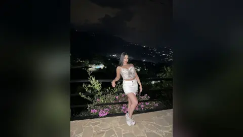 Young woman in a white lace crop top and denim skirt posing on a balcony at night overlooking city lights.