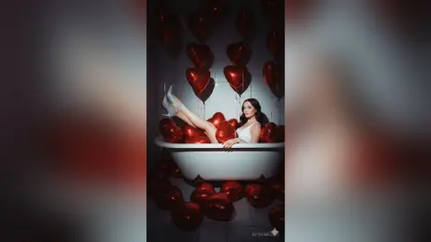 Glamorous woman in white bodysuit relaxing in a bathtub surrounded by red heart balloons in a moody setting.