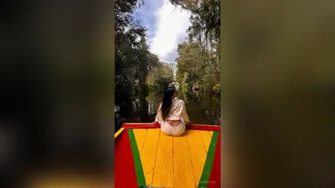 Woman in a white top sitting on a colorful boat on a peaceful river surrounded by lush green trees.