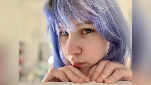 Close-up portrait of a young woman with lavender purple hair looking thoughtfully at the camera in soft lighting.