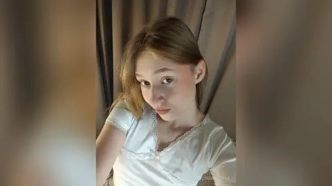 EvelinaMoorina looks into the camera with a soft expression, wearing a white lace blouse against a dark curtain background.