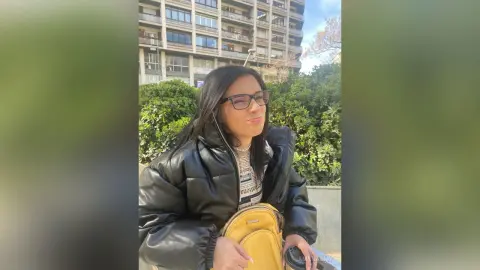 Smiling woman in a black puffer jacket and glasses standing outdoors near green bushes and city buildings.