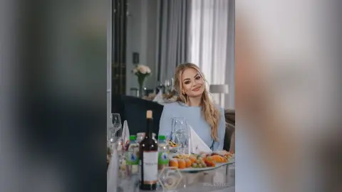 AshlieBridge smiles while sitting at a table with wine, glasses, and a plate of fruit in a bright room.