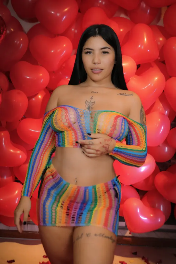 channeljonson — Young woman in colorful rainbow crochet crop top and mini skirt posing against a red heart balloon backdrop.