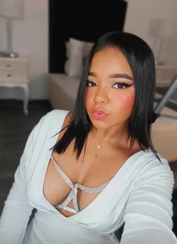 Young woman with long dark hair wearing a white low-cut top and gold necklaces in a cozy bedroom setting.
