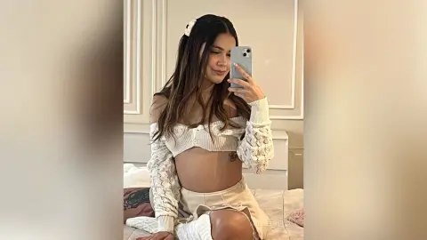 Smiling young woman in a white lace crop top and skirt sitting on a bed in a bright bedroom.