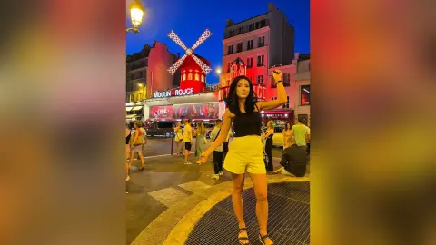 Woman in black top and yellow shorts posing at night in front of the illuminated Moulin Rouge in Paris.
