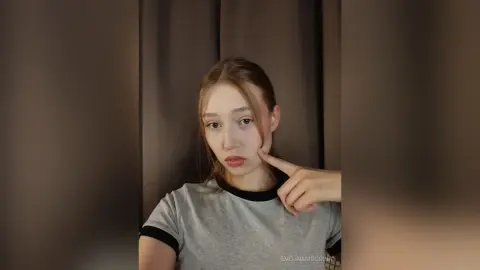 Young woman in a grey t-shirt with black trim posing thoughtfully against a dark curtain background.