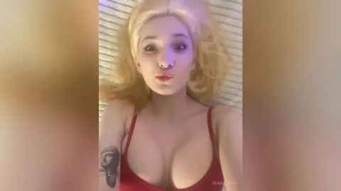 Blonde woman in a red tank top making a playful kissy face while relaxing on a white bed.