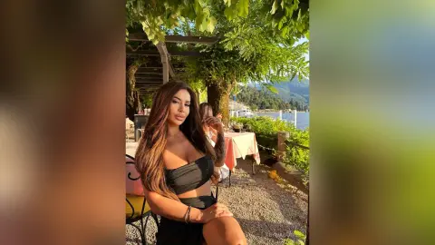 ReynaGomes poses outdoors in a black tube top, showing off her cleavage and long brown hair while sitting at an outdoor table near a lake.