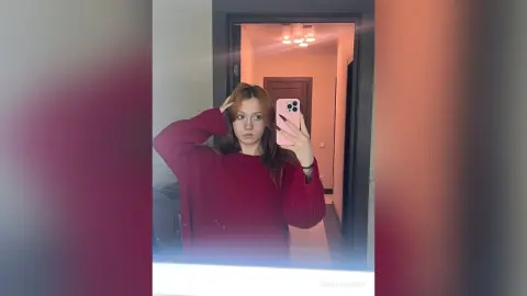 Young woman in a cozy red sweater taking a mirror selfie in a modern minimalist hallway.