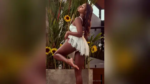 Elegant woman in a white lace bodysuit posing amidst tropical plants and sunflowers in a sunny outdoor setting.