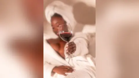 Relaxed Black woman in a white bathrobe enjoying a glass of red wine in a cozy, serene setting.