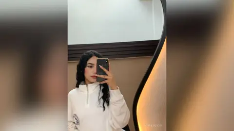 ZoeMonrow poses for a mirror selfie wearing a white long-sleeve zip-up top with her long dark hair down.