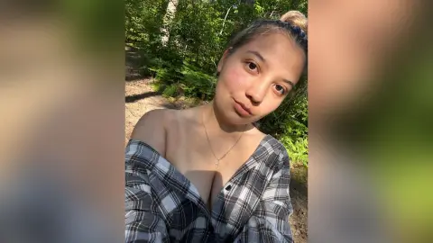 Young woman in a plaid shirt smiling during a sunny outdoor walk through a lush green forest.