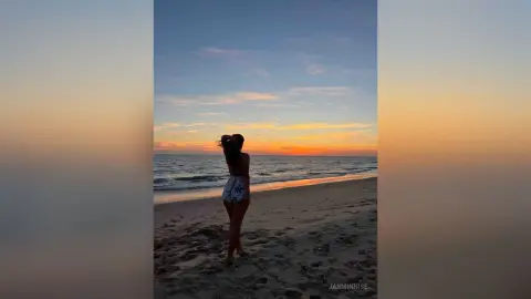JasminNise stands on a sandy beach at sunset, looking back over her shoulder while wearing a white patterned bikini or two-piece swimsuit.