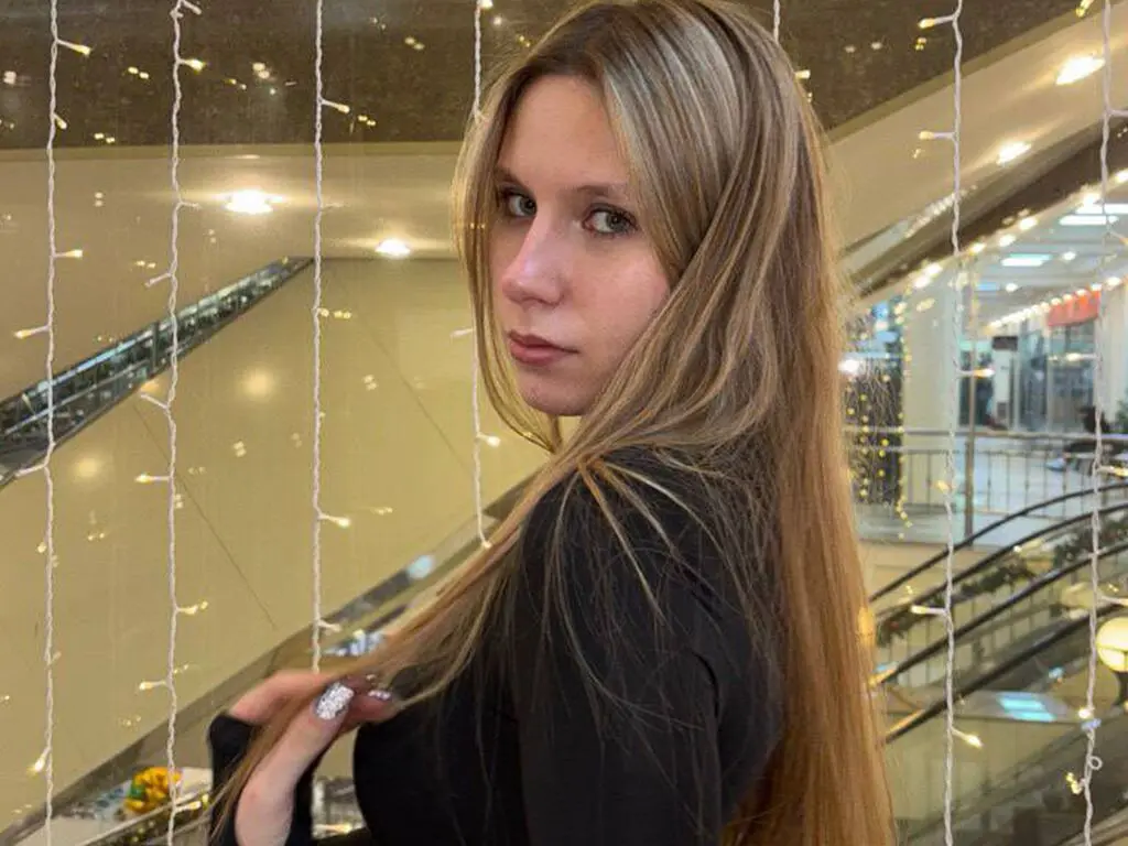 LydaCentorino looks over her shoulder with long blonde hair, wearing a black top in a bright indoor setting with hanging string lights.