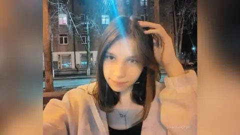 Young woman with long brown hair wearing a light pink jacket in a nighttime urban setting.