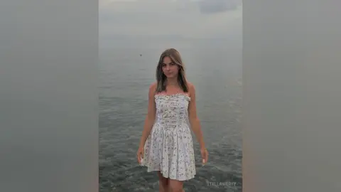 Young woman in a white floral sundress standing by the ocean under a cloudy sky.