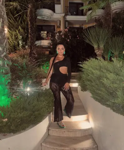 Stylish woman in a black cutout bodysuit and lace trousers posing on outdoor stairs at a tropical resort at night.