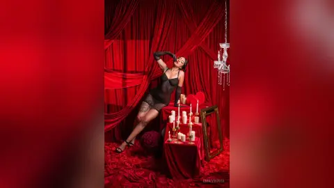 Glamorous woman in black lingerie posing against red velvet curtains with romantic candlelight decor.