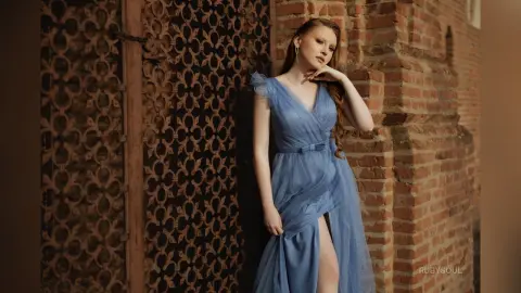 RubySoul poses in a light blue sheer dress with a high slit, standing against a brick wall and decorative metal gate.