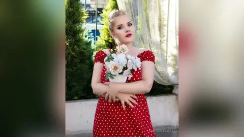 Blonde woman in a red polka dot dress holding a white flower bouquet on a sunny balcony.
