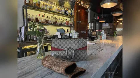 BossyBella — BossyBella's bag and a small brown pouch sit on a marble bar counter in a dimly lit bar setting with liquor bottles on shelves behind a glass vase.