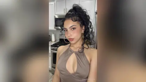 Young woman with curly hair wearing a taupe halter neck top in a modern kitchen setting.