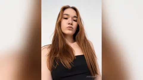 JaneBarbrick poses looking into the camera with long brown hair and a black top against a white background.