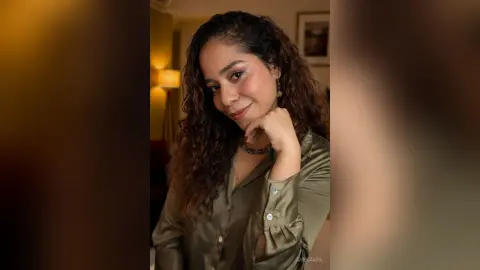Smiling woman with curly hair wearing an olive green silk blouse in a warm, cozy indoor setting.