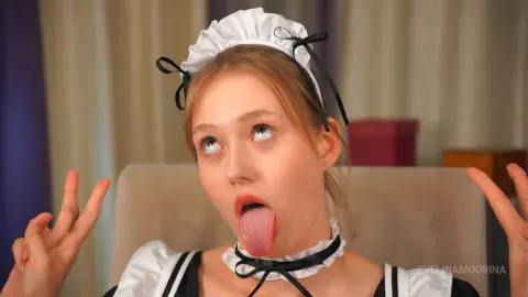 Playful blonde woman in a white maid costume sticking her tongue out in a cozy indoor setting.