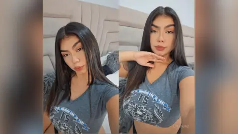 Young woman with long dark hair wearing a grey graphic crop top in a cozy bedroom setting.