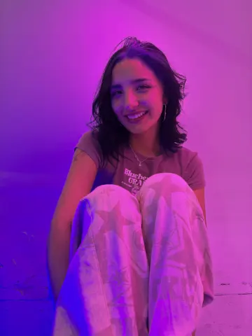 Smiling young woman in a graphic t-shirt and patterned lounge pants sitting under dreamy pink neon lighting.
