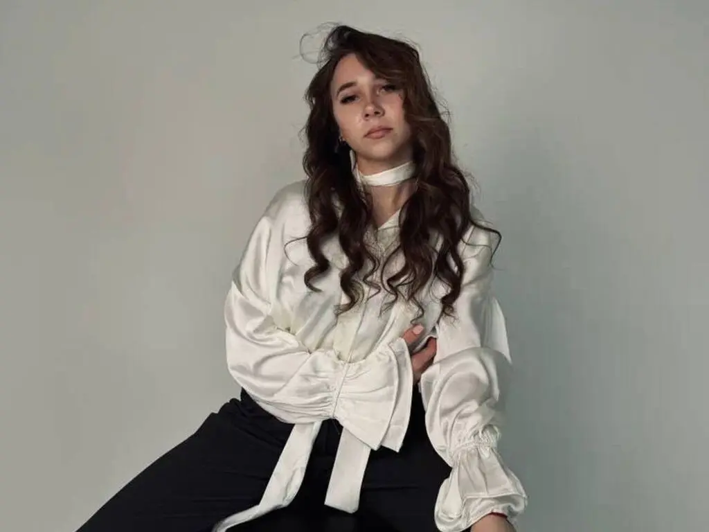 Young woman in a white satin blouse and black trousers posing against a neutral studio background.
