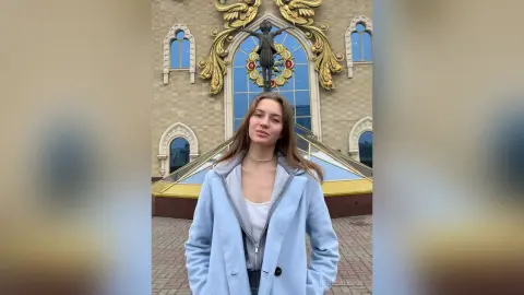 Young woman in a light blue coat standing before a grand gold-accented building on a bright day.