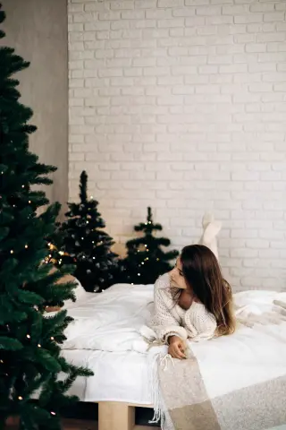 Cozy woman in a white knit sweater relaxing on a bed next to glowing Christmas trees in a bright room.