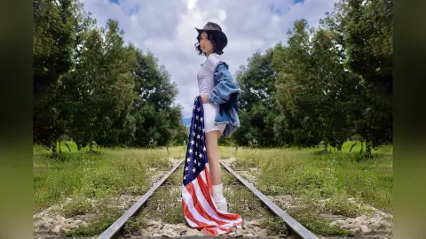 Fashionable woman in a white bodysuit and denim jacket holding an American flag on rustic railroad tracks.
