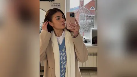 CharityRobes takes a mirror selfie wearing a tan coat, a white scarf, and a blue patterned sweater vest.