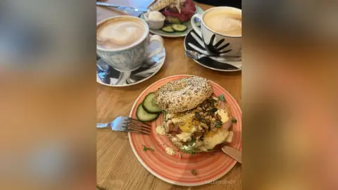 ValerieGracy's breakfast or brunch consisting of a seeded bagel topped with eggs and vegetables, served with a latte and cucumber slices.