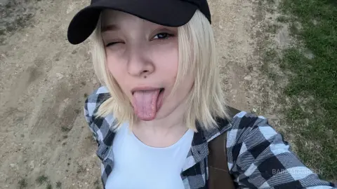 BarbaraGreen winks and sticks her tongue out, wearing a black cap and a plaid shirt while standing outdoors on a dirt path.