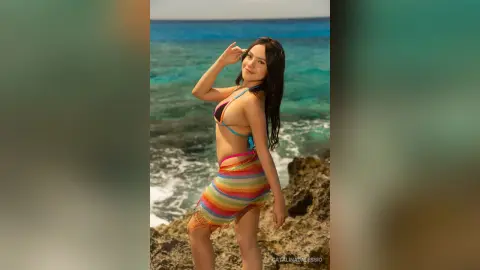 Smiling young woman in a colorful crochet bikini and sarong posing on a sunny tropical beach by the ocean.
