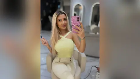 Anita poses for a mirror selfie wearing a yellow criss-cross halter top and light tan trousers.