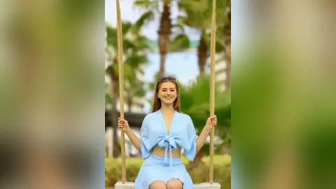 Smiling woman in a light blue summer outfit sitting on a swing in a sunny tropical garden setting.