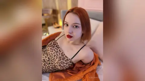 Redheaded woman in a leopard print top and orange cardigan relaxing in a cozy bedroom setting.