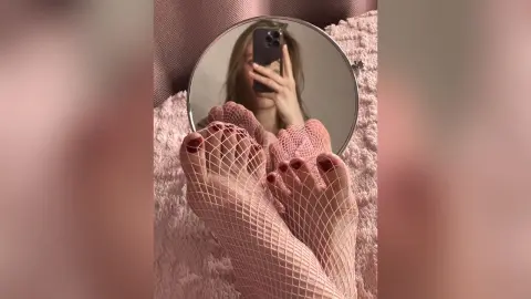 Woman wearing pink fishnet stockings sitting on a soft pink textured bed in a cozy, feminine setting.