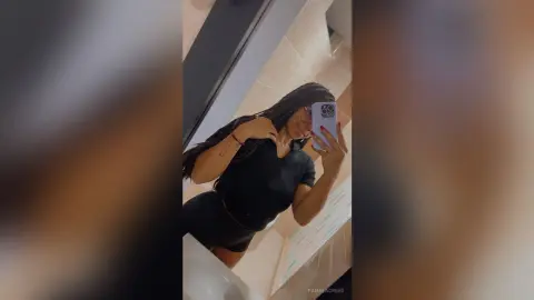 PamelaCreed poses in a black short-sleeve shirt and black shorts, taking a mirror selfie with her long dark hair down.
