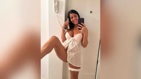 Smiling woman in white lace lingerie taking a mirror selfie in a bright modern bathroom.