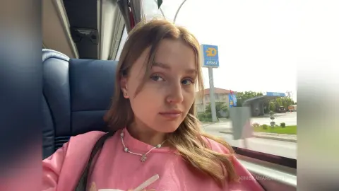 Young woman in a pink sweatshirt sitting on a bus looking out the window on a bright day.