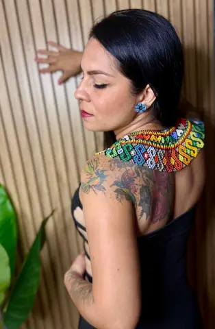 Woman with shoulder tattoos wearing a colorful beaded collar and black dress against a wooden wall.